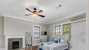 A bedroom with a white ceiling fan, a fireplace, a bed with floral bedding, and two windows with shutters. - Royal Resort (Jackson)