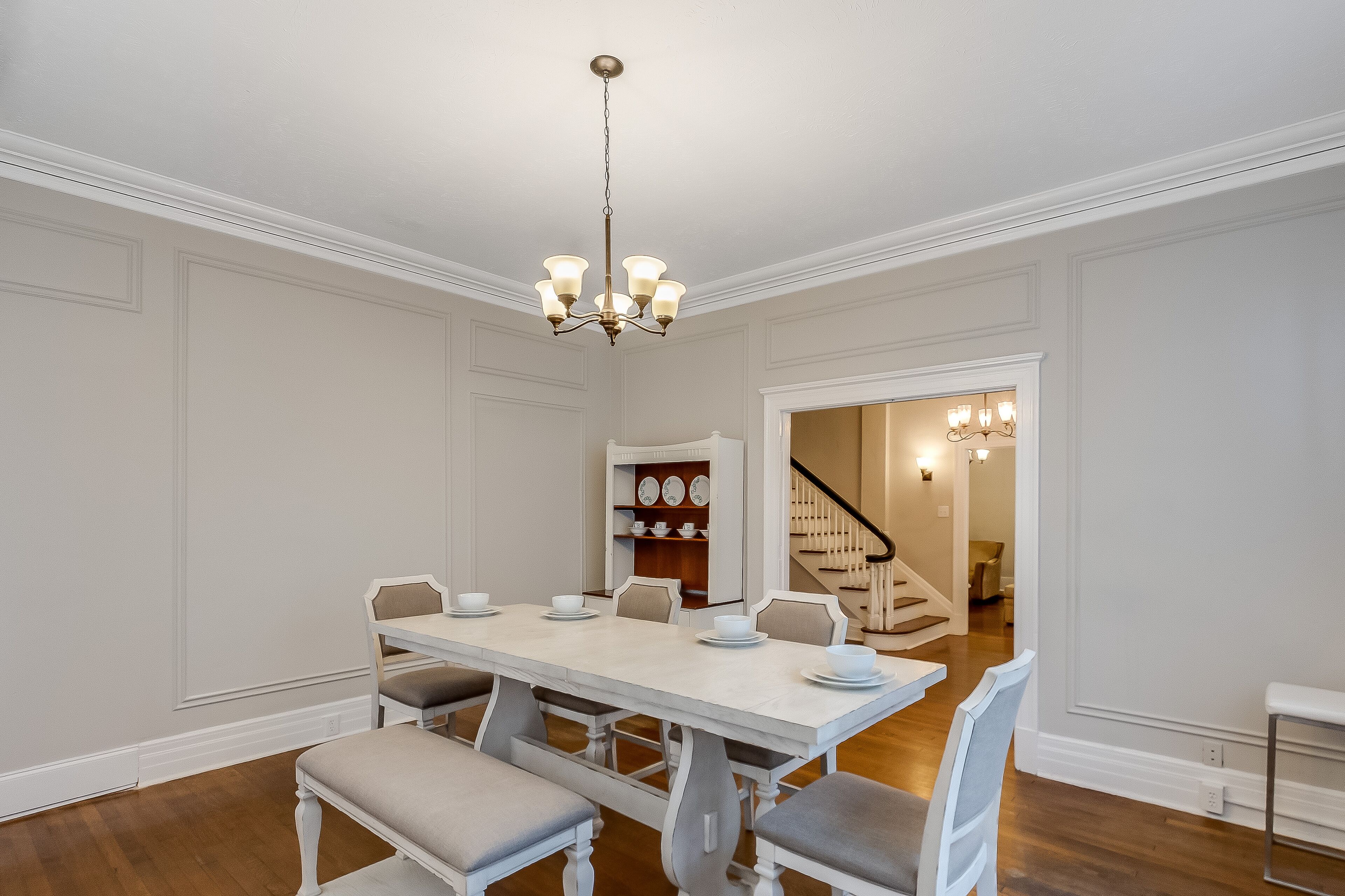 A dining room with a white table set for six, a chandelier, and a staircase leading to another room.