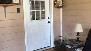 A room with a white door, wooden flooring, a black chair, a small table with a lamp, and a decorative wall hanging. - Although a country setting, the cottage has all modern conveniences & comforts (Climax)