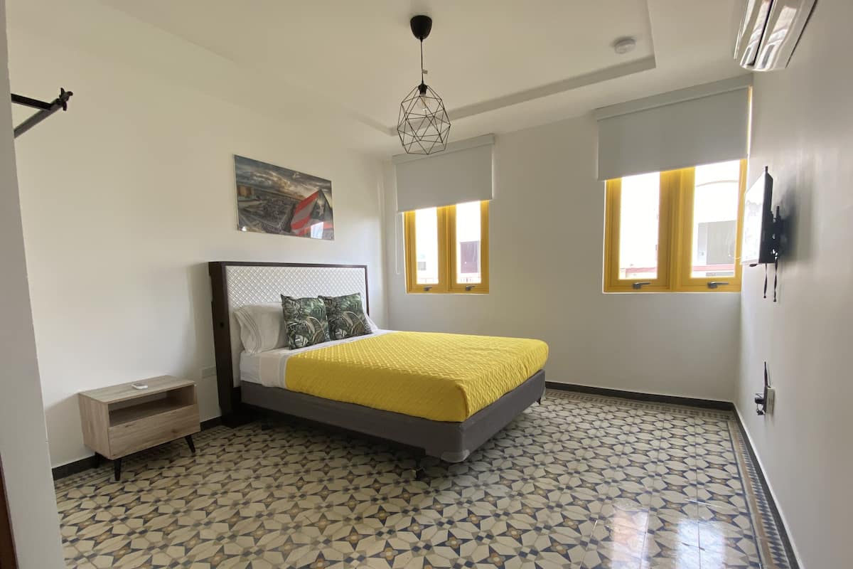 Une chambre à coucher comprenant un lit, une table de chevet, un tableau, un sol à motifs géométriques et un luminaire au plafond.