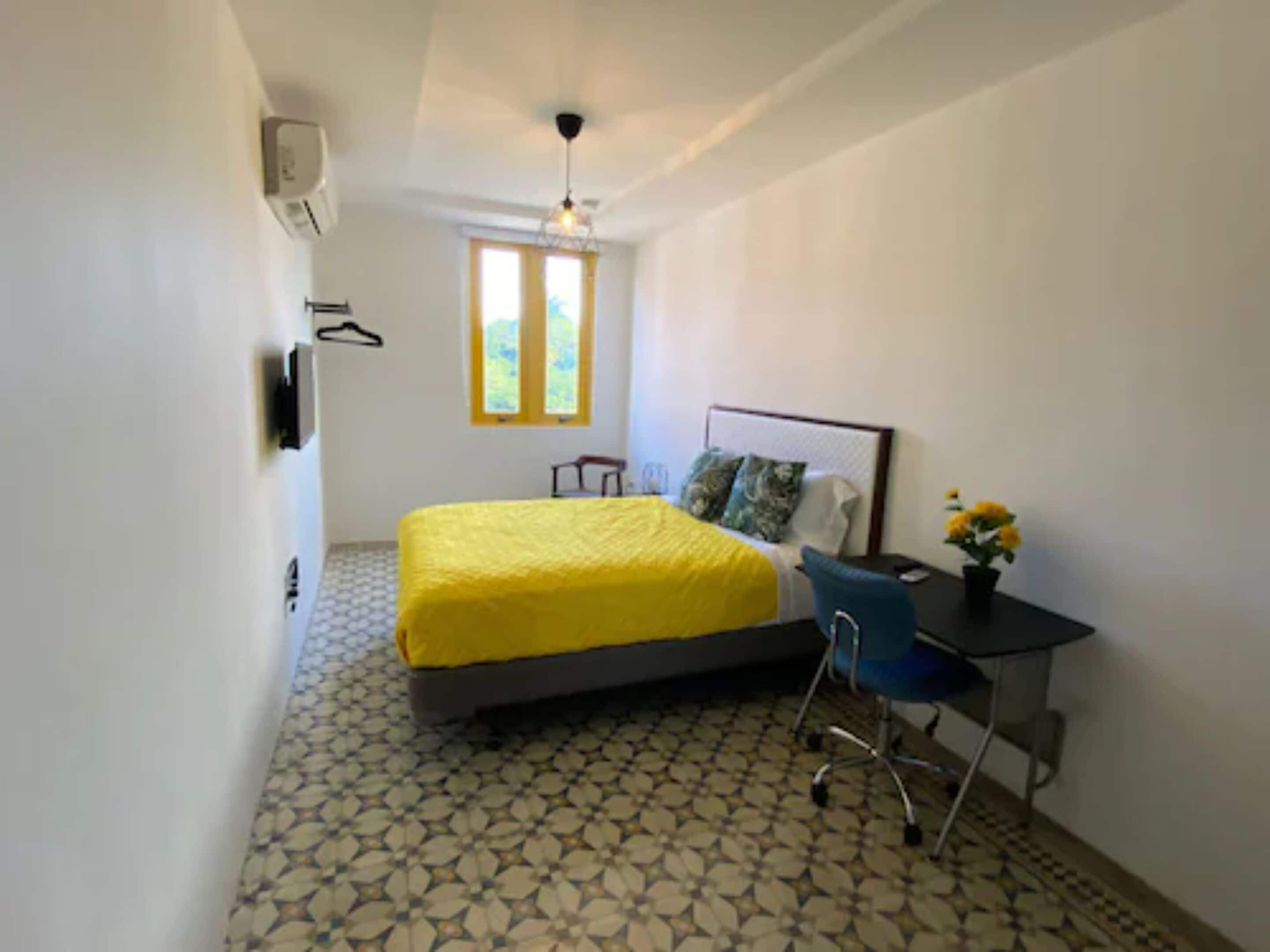 Un dormitorio con cama, escritorio, silla, ventana y aire acondicionado de pared.