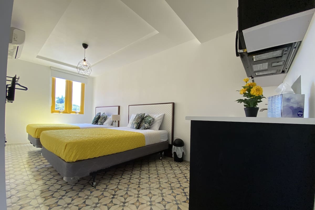 Une chambre d’hôtel avec deux lits, un sol carrelé, une fenêtre avec des rideaux jaunes, un luminaire au plafond et une petite plante en pot.