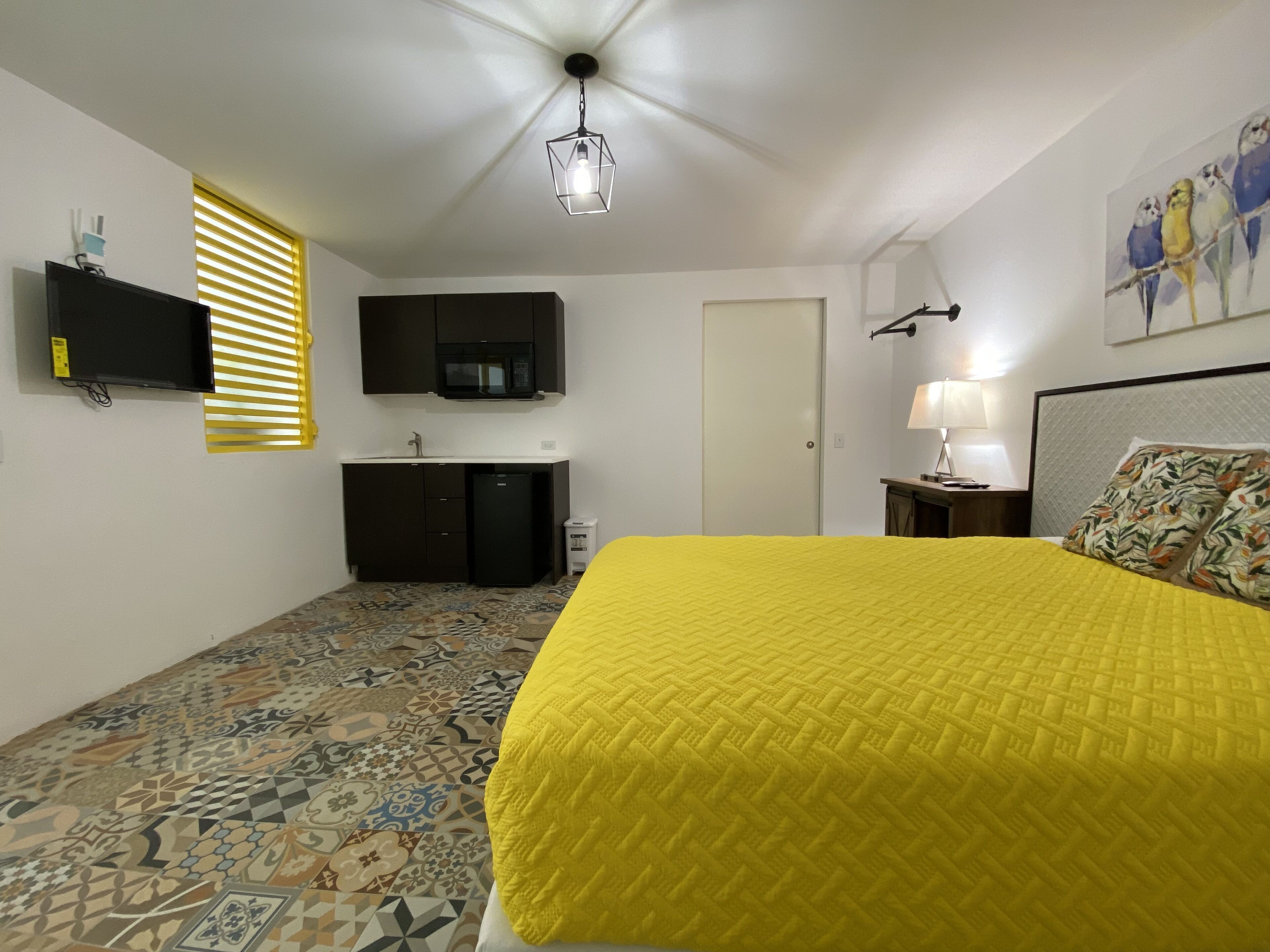 Camera da letto con pavimento a motivi, un letto con copriletto giallo, una TV a schermo piatto, un forno a microonde, un piccolo frigorifero, una lampada e un quadro appeso al muro.