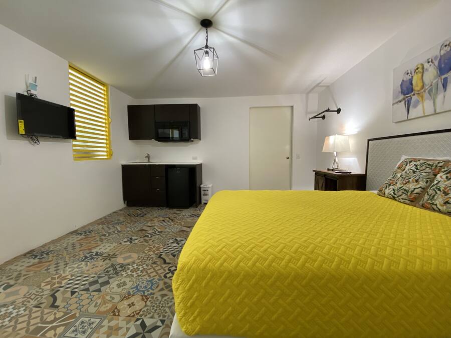 A bedroom with a patterned floor, a bed with a yellow bedspread, a flat-screen TV, a microwave, a small refrigerator, a lamp, and a painting on the wall.