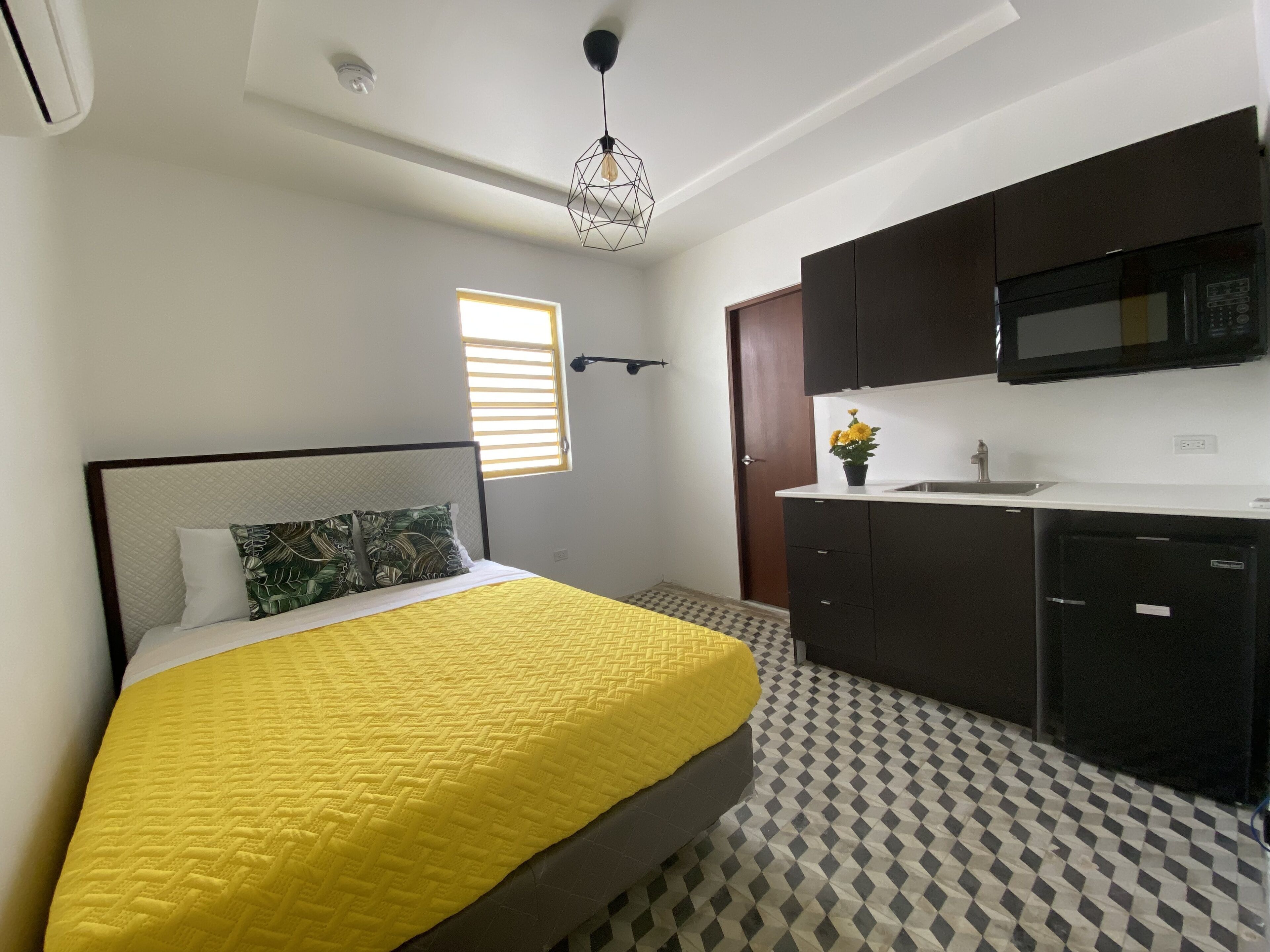 A bedroom with a bed, a window, a kitchen area with a microwave, and a patterned floor.