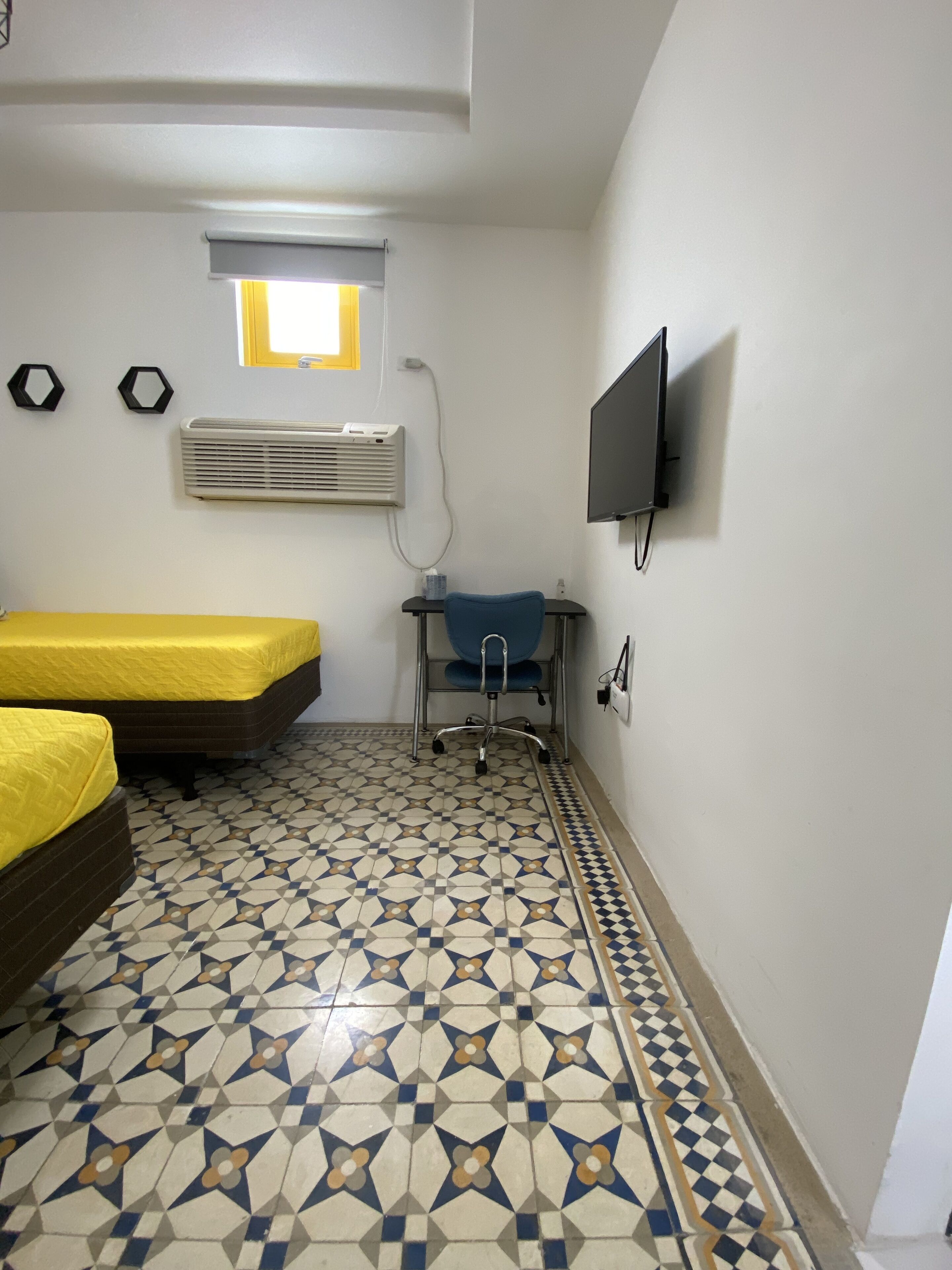 Una habitación con dos camas, un escritorio con silla, un televisor fijado a la pared, aire acondicionado y un suelo con diseño.