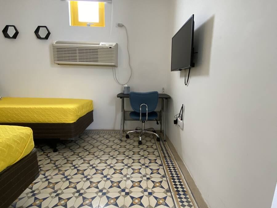 Una habitación con dos camas, un escritorio con silla, un televisor fijado a la pared, aire acondicionado y un suelo con diseño.