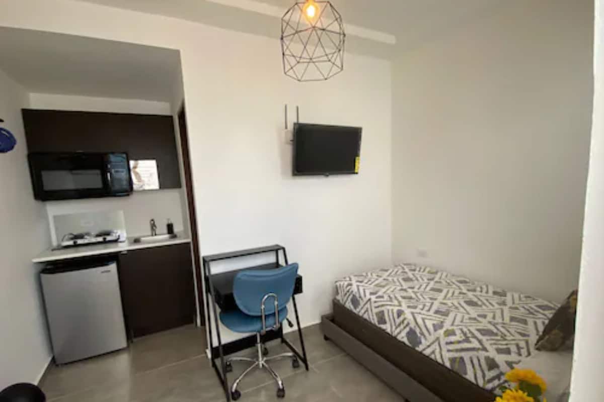 Un espace de vie compact comprenant un coin cuisine, un lit avec une literie à motifs, une chaise bleue et un téléviseur fixé au mur.