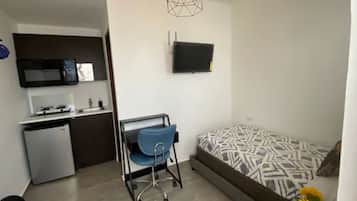 A compact living space with a kitchenette, a bed with patterned bedding, a blue chair, and a TV on the wall.
