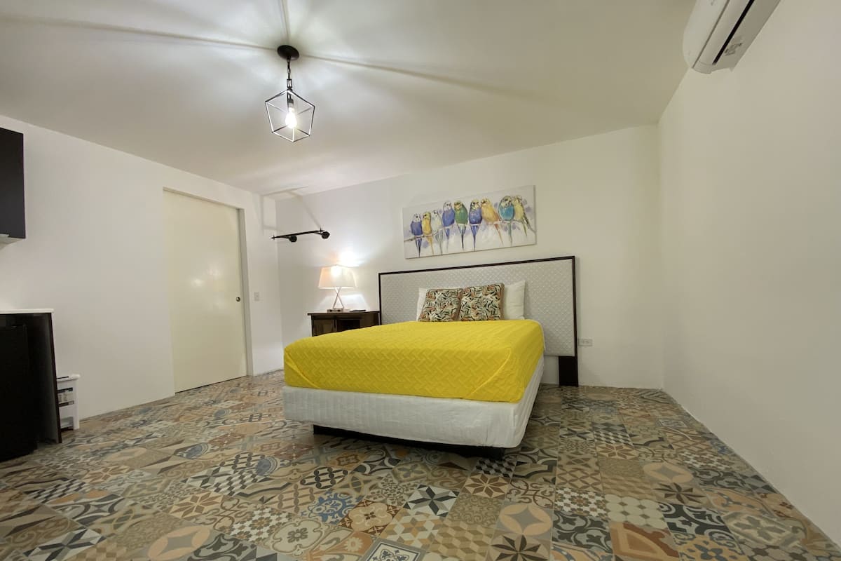 Une chambre avec un sol à motifs, un lit recouvert d’une couverture jaune, un téléviseur fixé au mur et un ventilateur de plafond.