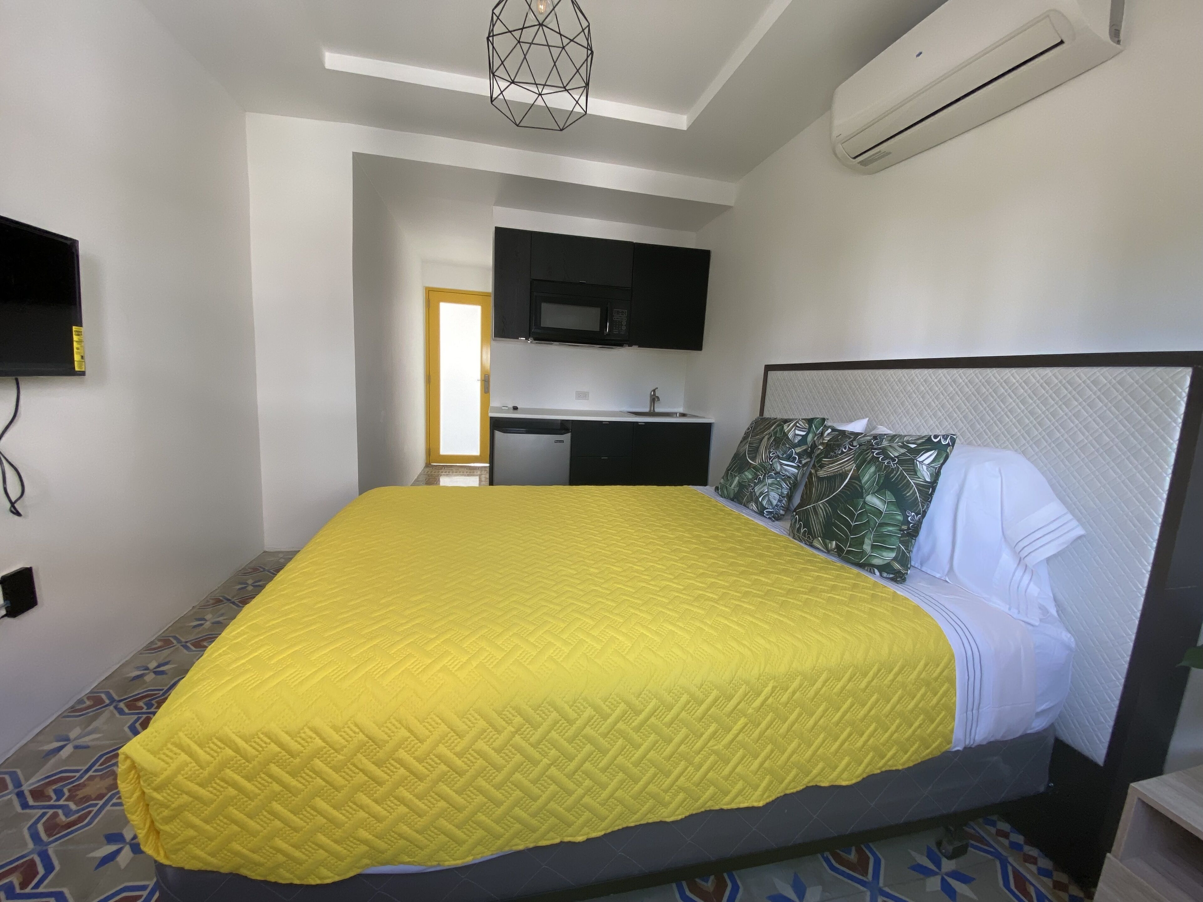 A hotel room with a bed, a small kitchenette, a TV, and an air conditioning unit.