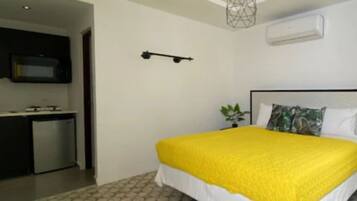 A bedroom with a bed, a bedside table, a small plant, and a kitchen area with a microwave.