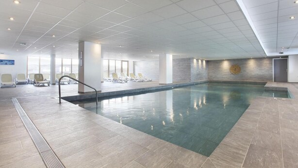 Ein großes Hallenbad mit Fliesenboden, einer Metallleiter und mehreren Liegestühlen rund um den Pool.