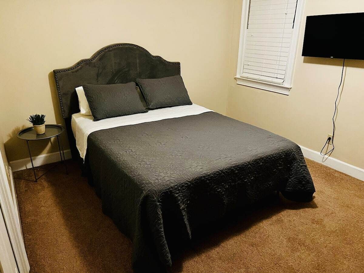 Una cama bien hecha con un cabecero oscuro y almohadas, una mesita de noche con una planta y un televisor montado en la pared.