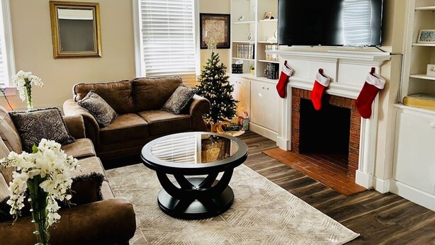 A cozy living room with a brown leather sofa, a glass coffee table, a fireplace with stockings, and a TV on the wall.