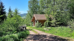A rustic wooden cabin with a tiled roof, surrounded by lush greenery and a clear sky.