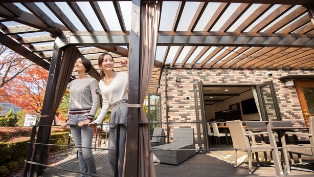 A covered outdoor area with a wooden deck, outdoor seating, and a brick building in the background. - HOTORI no TERRACE BAN (Fujikawaguchiko)