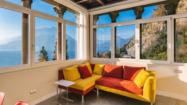 Ein Zimmer mit einem gelb-roten Ecksofa, einem kleinen weißen Tisch und großen Fenstern mit Blick auf Berge und Meer.