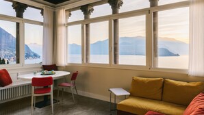 Ein Zimmer mit einem großen Fenster, das Blick auf Berge und einen See bietet. Es gibt einen weißen Tisch mit Stühlen, ein gelbes Sofa und einen Heizkörper unter dem Fenster.