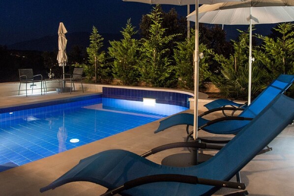 A swimming pool with blue tiles, surrounded by lounge chairs and umbrellas, with trees in the background.