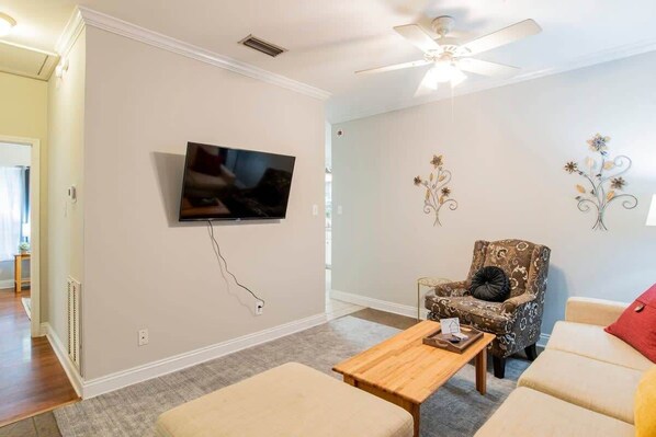 A living room with a flat-screen TV mounted on the wall, a patterned armchair, a wooden coffee table, and a beige sofa.
