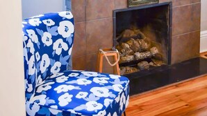 Een woonkamer met een open haard, een blauwe fauteuil met bloemenmotief en twee vazen met witte bloemen.