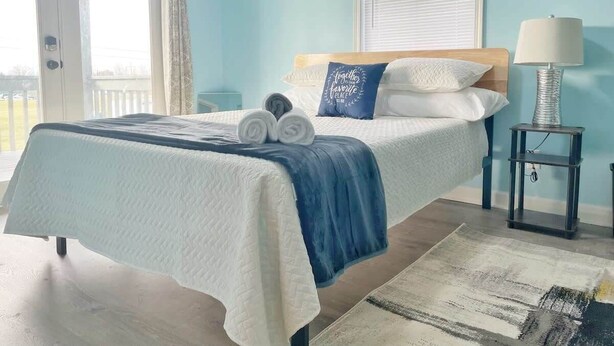 A bedroom with a bed, a nightstand, a lamp, and a view through a window. - The Bliss Lodge - Large House (Columbus)