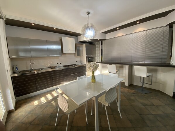 A modern kitchen with a white dining table, dark cabinetry, and a large window with blinds.