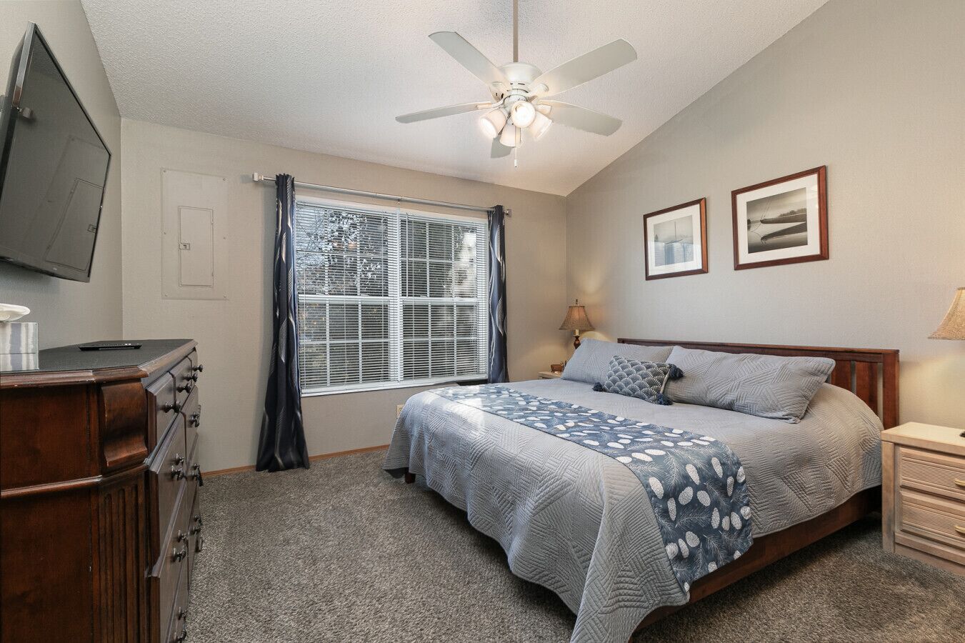 Une chambre à coucher comprenant un lit, un ventilateur de plafond, une fenêtre avec des stores, une commode et un téléviseur fixé au mur.