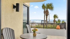 A balcony with a table and chairs, a vase with flowers, and a view of the ocean and palm trees.