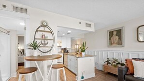 A modern kitchen with white cabinetry, a central island, and bar stools. There is a dining area with a round table and two bar stools. The kitchen features a built-in stove, a sink, and a refrigerator. The space is decorated with plants and a painting on the wall.