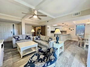 A spacious living area with a sofa, coffee table, and a dining table. A ceiling fan and kitchen are visible in the background.