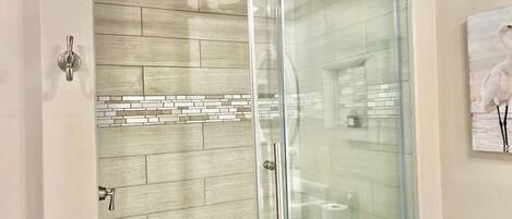 A glass-enclosed shower with a rain showerhead, a tiled wall, and a toilet with a toilet paper dispenser.