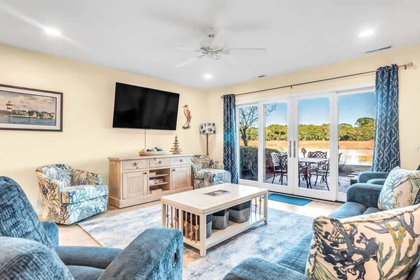 A living room with a flat-screen TV, a sofa, a coffee table, and a view through a glass door to an outdoor patio.