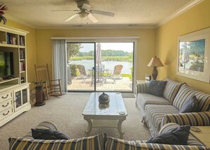 A living room with a sofa, a coffee table, a bookshelf, a television, and a view of a lake through a sliding glass door.