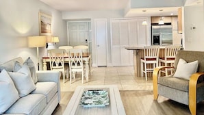 A modern living room with a sofa, a dining table with chairs, a kitchen area with a refrigerator, and a door leading to a closet.