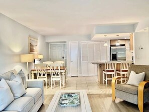 A modern living room with a sofa, a dining table with chairs, a kitchen area with a refrigerator, and a door leading to a closet.