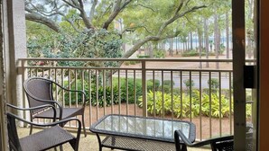 A balcony with two chairs and a table, surrounded by a railing, overlooking a landscaped garden and a road.