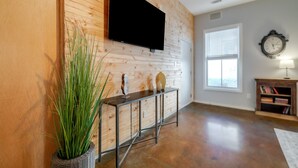 A modern living room with a wooden wall, a flat-screen TV mounted above a console table, a large window with blinds, and a clock on the wall. - ⭐Southern Charmer•Historic Downtown•Private Porch⭐ (Savannah)