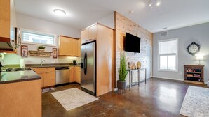 A modern kitchen with wooden cabinets, a stainless steel refrigerator, and a flat-screen TV mounted on a brick wall. - ⭐Southern Charmer•Historic Downtown•Private Porch⭐ (Savannah)