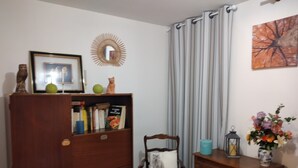 Ein Zimmer mit einem Holzkasten, einem Stuhl mit Sitzkissen, einem kleinen Tisch mit Blumenvase sowie einer Wand mit Spiegel und Gemälde.
