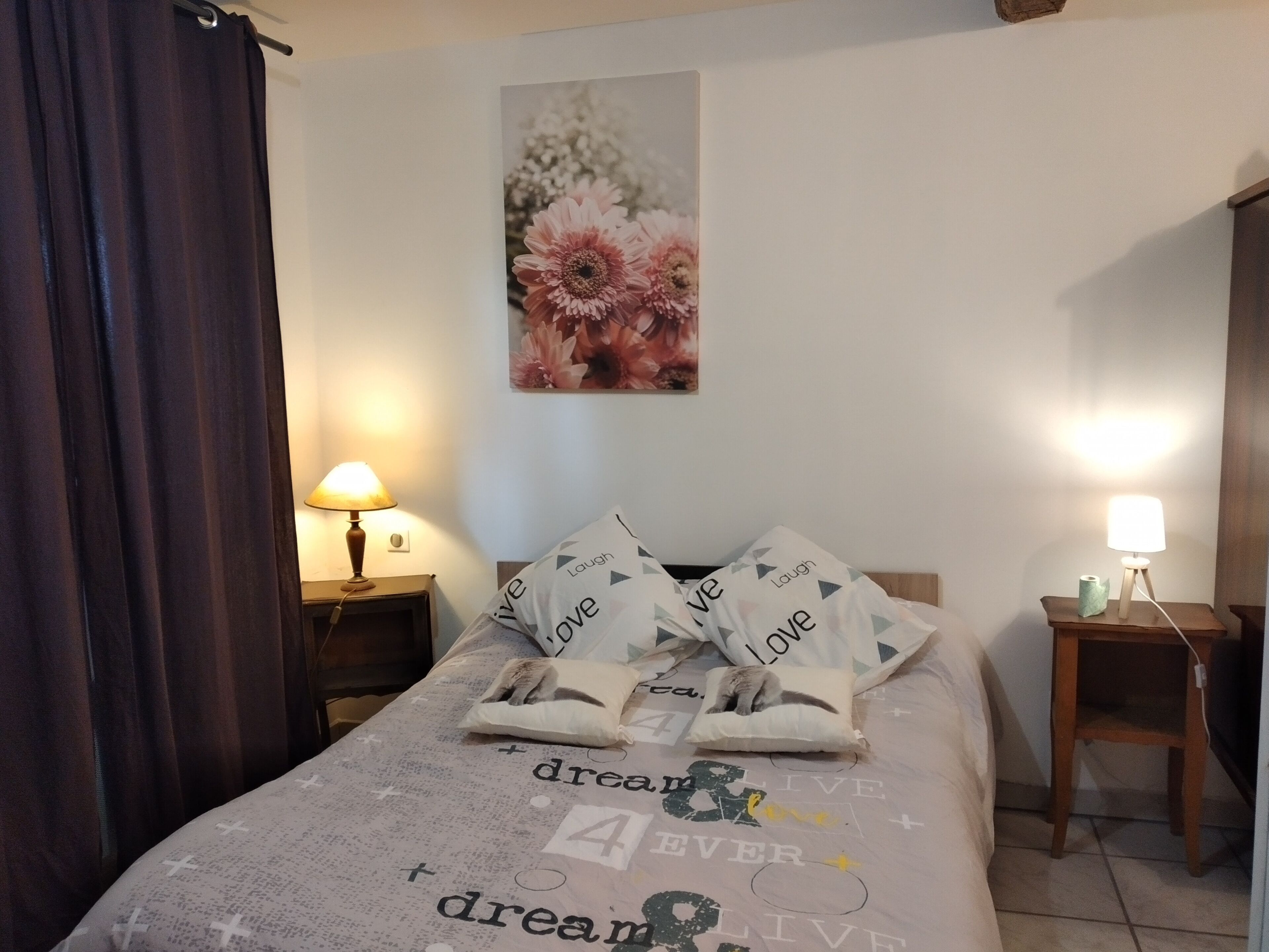 A neatly made bed with a motivational print, two bedside lamps, a wall-mounted flower painting, and a dark curtain.