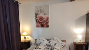 Ein ordentlich bezogenes Bett mit floralem Bettbezug und Kissen mit dem Schriftzug "love". Zwei Nachttische mit Lampen und ein Wandbild über dem Bett.
