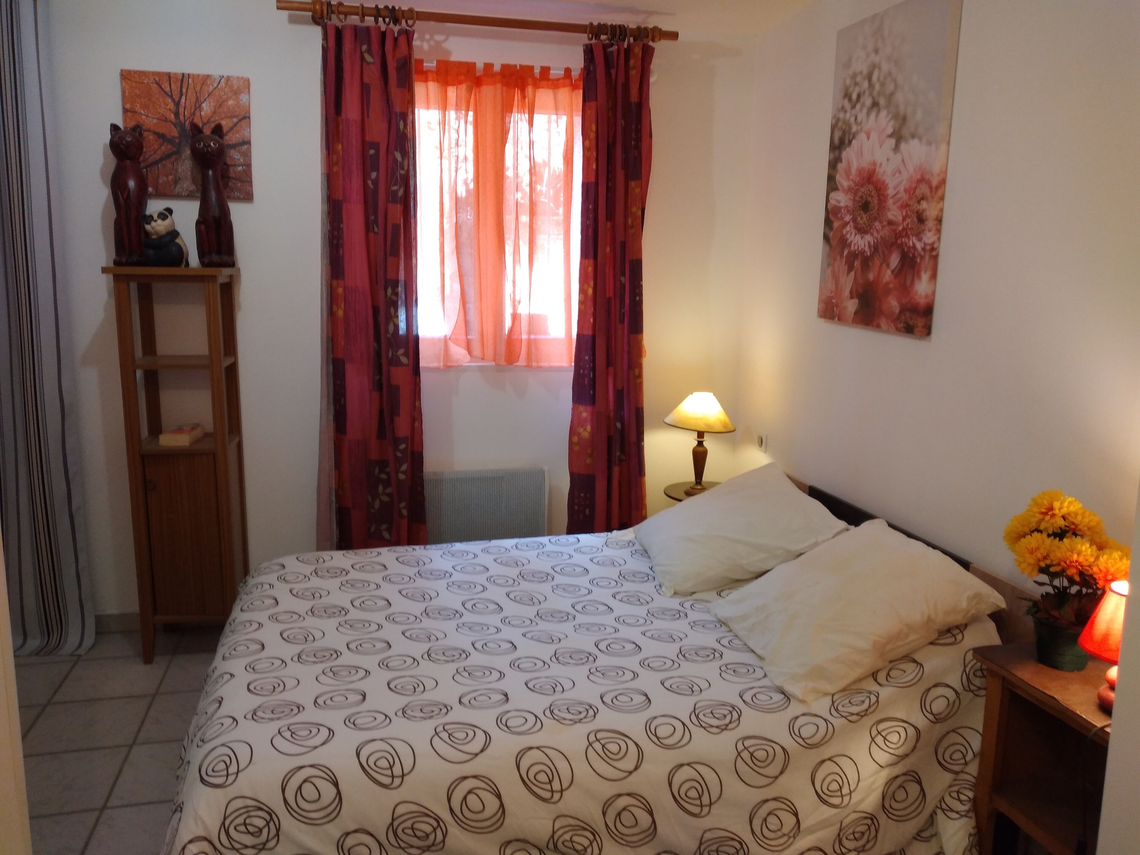 A bedroom with a bed, a nightstand with a lamp, a shelf with decorative items, and a window with curtains.