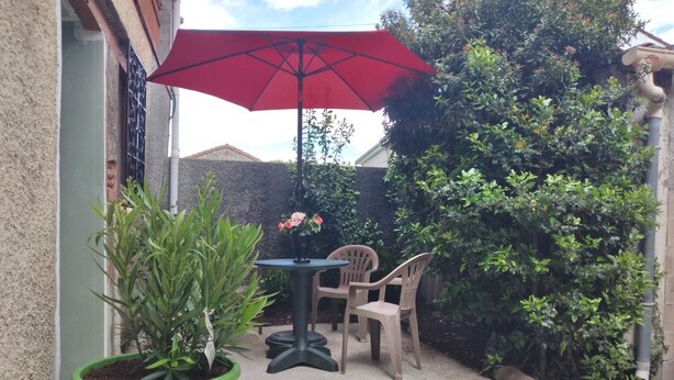 Eine Terrasse mit einem grünen Topfpflanzen, einem Tisch für zwei und einem roten Sonnenschirm.
