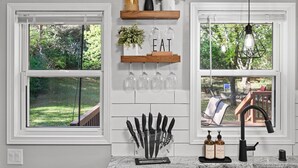 Une cuisine avec des meubles blancs, une étagère en bois où sont rangés des verres, un bloc à couteaux et un luminaire suspendu.