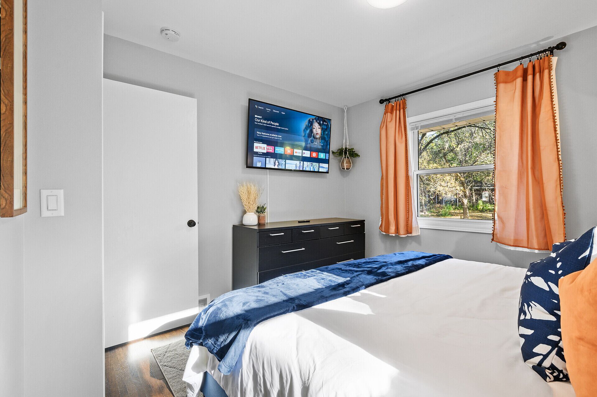 Une chambre à coucher avec un lit, une commode, un téléviseur fixé au mur et une fenêtre avec des rideaux.