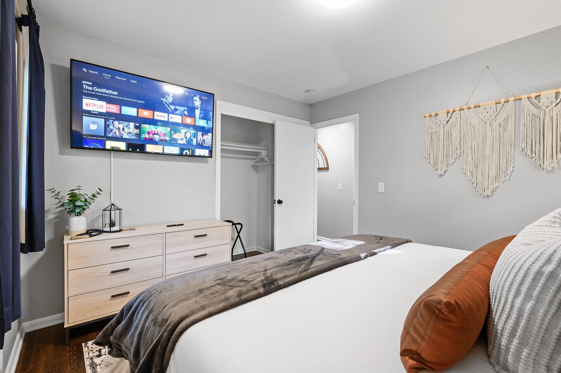 A modern bedroom with a bed, a TV mounted on the wall displaying a streaming service, a dresser, and a macrame wall hanging.