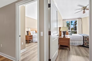 Quarto de hotel com cama, mesa de cabeceira, abajur e vista para palmeiras pela janela.