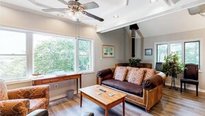 Sala de estar com sofá de couro marrom, mesa de centro de madeira e ventilador de teto. Janelas amplas oferecem luz natural e vista para árvores.